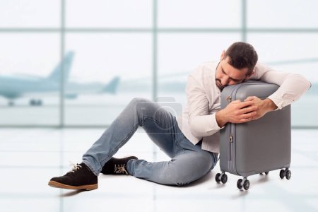 Foto de Hombre barbudo sentado en el suelo y abrazando su bolso de viaje dormido y cansado de esperar en un aeropuerto. Largas colas de espera. Retrasos de vuelo. Desesperación por vuelos cancelados - Imagen libre de derechos
