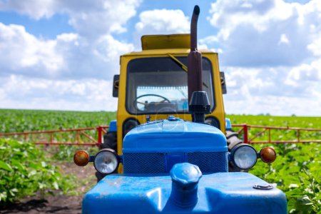 Photo for An old farm tractor painted in yellow-blue color with a trailed sprayer with tank capacities is preparing to spray a crop protection product on a field sown with sunflowers. - Royalty Free Image