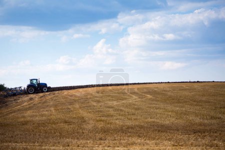 Foto de Un moderno tractor azul con un arado reversible unido con un rodillo descascarillado está arando un campo en el que se acaba de cosechar el cultivo de grano de primavera. A mediados de verano en Ucrania central. - Imagen libre de derechos