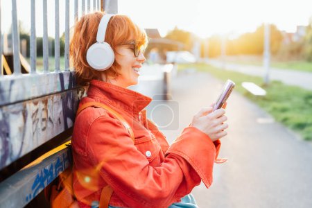 Photo for Smiling stylish woman in bright clothes and sunglasses wearing wireless headphones on her head seting phone for listening music. Walk in city park on sunset. Hipster lifestyle. Selective focus - Royalty Free Image