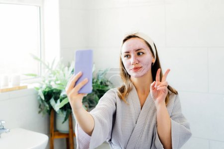 Téléchargez les photos : Femme souriante avec masque en argile rose facial prenant selfie avec téléphone mobile à la maison bain profitant de détente et de soins de beauté spa. Blogueur beauté créant du contenu. Mise au point sélective, espace de copie. - en image libre de droit