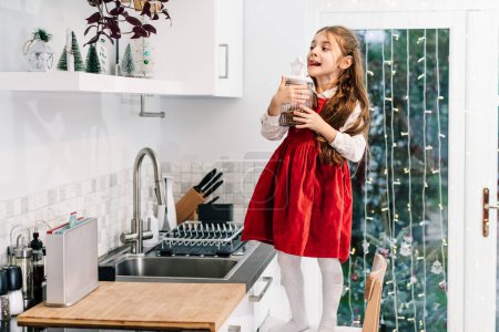 Photo for Happy smiling cute little girl kid standing on the chair and reached jar with chocolate festive cookies on the shelf on decorated for winter Christmas holidays modern kitchen. Overeating sweets. - Royalty Free Image
