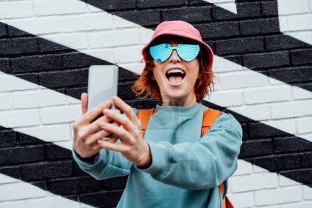 Photo for Excited redhead woman screaming while taking a selfie photo outdoors. Emotional hipster fashion women in bright clothes, heart shaped glasses, bucket hat taking selfie photo on the phone camera. - Royalty Free Image