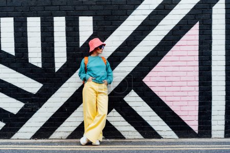 Photo for Hipster fashion young woman in bright clothes, sun glasses and bucket hat posing on the painted brick wall background. Urban city street fashion. Selective focus. Copy space. - Royalty Free Image