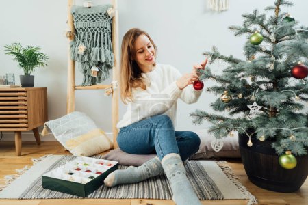 Young woman in cozy sweater decorating potted Christmas tree with small glass baubles in light modern Scandinavian interior. Eco-friendly winter holidays. Christmas tree in a pot. Selective focus.
