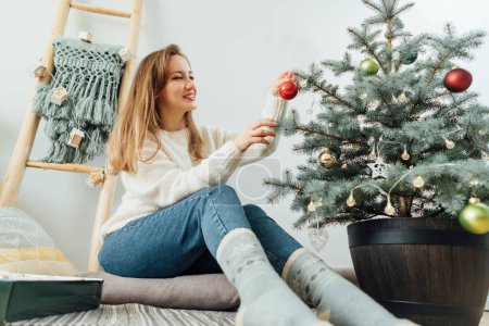 Téléchargez les photos : Jeune femme en pull confortable décorant l'arbre de Noël en pot avec de petites boules de verre à l'intérieur scandinave moderne léger. Vacances d'hiver écologiques. Arbre de Noël dans un pot. Concentration sélective. - en image libre de droit