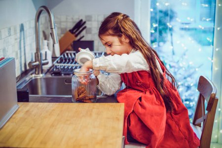 Photo for Happy smiling cute little girl kid standing on the chair and reached jar with chocolate festive cookies on the shelf on decorated for winter Christmas holidays modern kitchen. Overeating sweets - Royalty Free Image