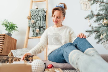 Photo for Young woman in cozy sweater petting her cat while decorating potted Christmas tree in light modern Scandinavian interior home. Eco-friendly winter holidays. Domestic pet at home. Selective focus - Royalty Free Image