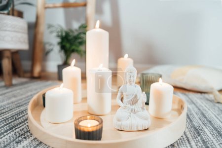 Wooden tray with burning candles and white Buddha statuette on the floor of modern Scandi interior. Zen Composition for meditation, yoga practice, relaxation. Balance and calm energy flow indoor