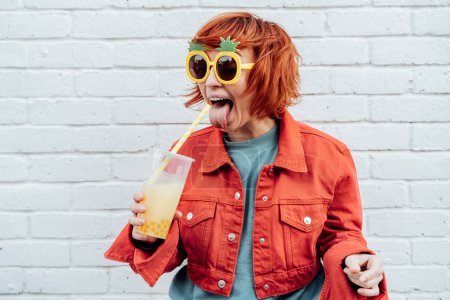 Photo for Funny Hipster fashion young woman protruding tongue in bright clothes and pineapple glasses drinking fruity tapioca bubble tea with straw on the white brick wall background. Selective focus - Royalty Free Image