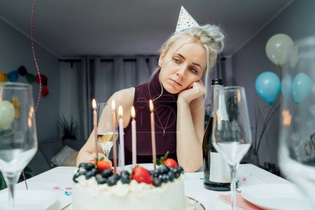Bored, sad attractive woman drinking champagne while celebrating birthday at home, sitting alone at served table with cake, keeping hand under chin, looking away, dreaming. Selfparty. Selective focus.