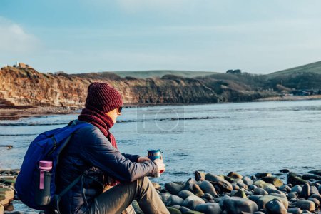 Téléchargez les photos : A young man in warm clothes sitting on the stone, drinking hot tea or coffee from reusable thermos cup and enjoying rocks and sea view. Relax during a walk on the coast. Cold season travel - en image libre de droit