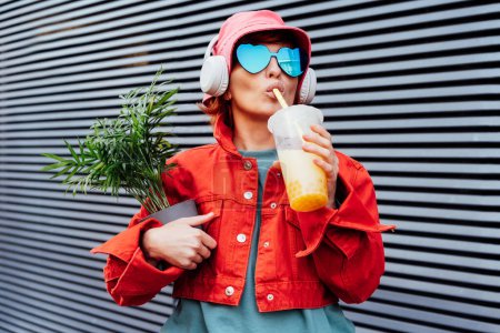 Foto de Hipster fashion woman in bright clothes, heart shaped glasses, headphones, bucket hat drinking fruity flavored tapioca bubble tea and holding green potted plant on the gray striped wall background - Imagen libre de derechos