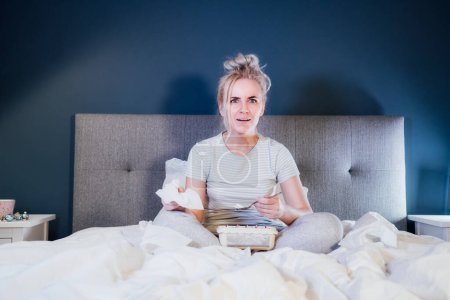 Photo for Young excited woman in home clothes passionately watching Tv or movie with bated breath. Girl eating ice cream, holding tissue while sitting on bed at home in evening alone. Leisure, relaxation time. - Royalty Free Image