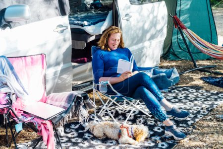 Téléchargez les photos : Adult woman relaxing, sitting in the travel chair and reading book on the background of camper car vehicle. Female living on camper car with animals and travel the world. Caravan car Vacation. - en image libre de droit