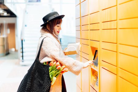 Photo for Fashion woman with box using modern postal automatic mail terminal with self service device for pickup or refund an order. Electronic locker for storing parcels. Online shopping. Selective focus. - Royalty Free Image