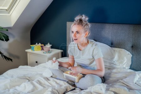 Photo for Young excited woman in home clothes passionately watching Tv or movie with bated breath. Girl eating ice cream, holding tissue while sitting on bed at home alone. Leisure, relaxation time. - Royalty Free Image