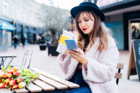 Photo for Stylish young woman enjoying coffee from a reusable cup, reading text message on mobile phone at coffee shop . Fresh tulips bouquet on the table. Springtime street fashion. Influencer. Selective focus - Royalty Free Image