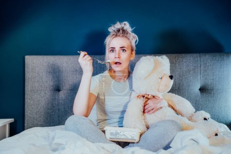 Photo for Young woman in home clothes passionately, hugging teddy bear, watching movie with bated breath. Girl eating ice cream while sitting on bed at home in evening alone. Leisure, relaxation time - Royalty Free Image