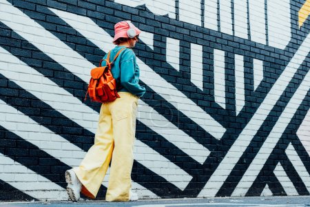 Back view stylish woman in bright clothes and bucket hat wearing wireless headphones and listening to music while walking near city urban painted wall. Fashionable hipster lifestyle. Selective focus