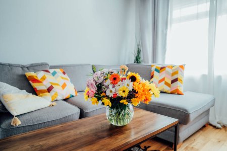 Vase with huge multicolor various flower bouquet on the coffee table with blurred background of modern cozy light living room with gray couch sofa. Giving flowers. Mothers day, birthday gift