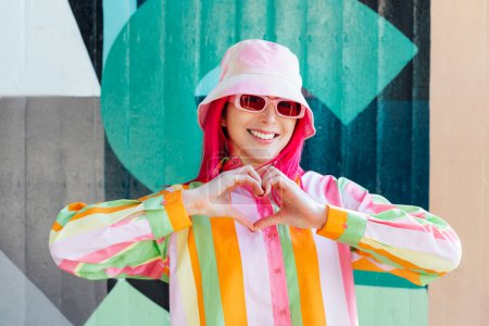 Photo for Vanilla Girl. Kawaii vibes. Candy colors design. Young woman with pink hair and sun glasses in bucket hat and multicolor shirt making sign Heart by fingers on wall background. Love fashion concept - Royalty Free Image