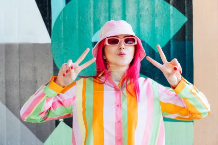 Photo for Vanilla Girl. Kawaii vibes. Candy colors design. Young woman with pink hair and sunglasses in Bucket hat and multicolor strippled shirt making V sign by fingers. Anime Urban street fashion - Royalty Free Image
