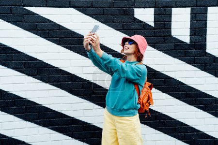 Téléchargez les photos : Femme rousse prenant une photo selfie avec sortir la langue à l'extérieur. Femme de mode hipster émotionnelle en vêtements lumineux, lunettes en forme de coeur, chapeau seau prenant selfie photo sur l'appareil photo - en image libre de droit