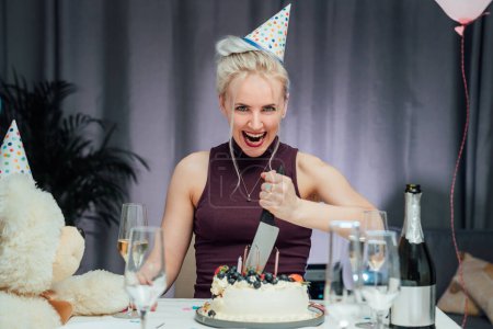 Foto de Atractiva loca y risueño cumpleañera con cuchillo de pan planeando cortar su pastel de cumpleaños mientras celebra su cumpleaños en casa sola. Enfoque selectivo. - Imagen libre de derechos