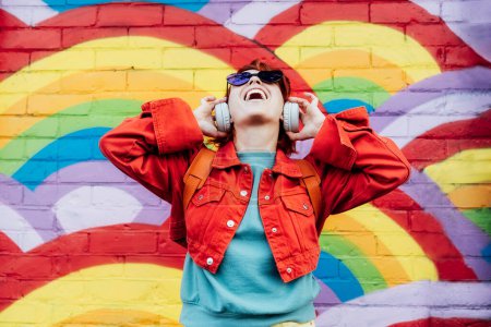 Foto de Emotional stylish woman in bright clothes wearing wireless headphones, listening to music and singing on rainbow graffity on the brick wall background. Fashionable hipster lifestyle - Imagen libre de derechos