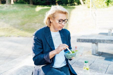 Téléchargez les photos : Femme d'affaires d'âge moyen mangeant de la salade de sa boîte à lunch assise sur le banc au parc du bureau. Boîte à lunch régime équilibré pour la perte de poids. Habitudes alimentaires saines et bien-être. Concentration sélective - en image libre de droit