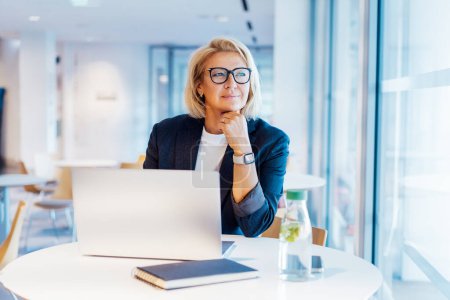 50s confident mature businesswoman dreamy looking at window, middle-aged experienced senior female professional working on laptop in open space office. Female entrepreneur working remotely