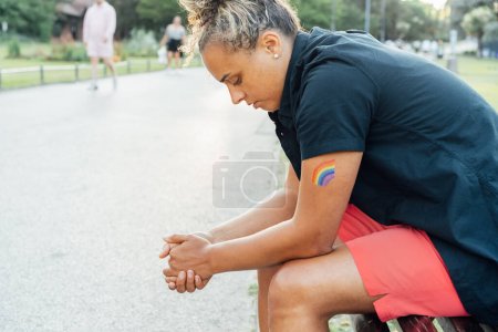 Sad frustrated lesbian woman with LGBTQ rainbow tattoo on her hand sitting on the park bench alone. Concept of loneliness, sadness and pain for homosexual discrimination. Mental crisis, depression.