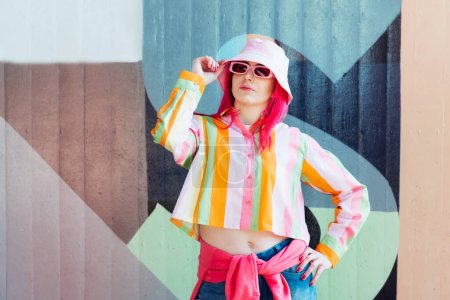 Photo for Urban Fresh street fashion look. Vanilla Girl. Kawaii vibes. Candy colors design. Bucket hat trends. Young woman with pink hair and sun glasses in multicolor strippled shirt on the wall background - Royalty Free Image
