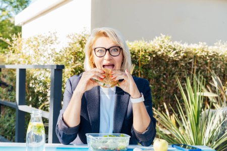 Téléchargez les photos : Portrait d'une femme d'affaires affamée et drôle d'âge moyen avec les yeux grands ouverts et la bouche mangeant un hamburger sain pendant le déjeuner au café en plein air du parc pendant sa pause. Habitudes alimentaires saines et bien-être - en image libre de droit