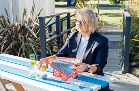 Téléchargez les photos : Femme d'affaires d'âge moyen sort son repas du midi au café extérieur du parc, aire de pique-nique pendant sa pause. Plats à emporter régime équilibré boîte à lunch avec salade. Habitudes alimentaires saines et bien-être. - en image libre de droit