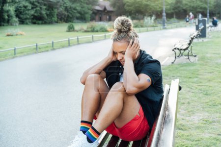 Sad lesbian woman with LGBTQ rainbow tattoo covering her head with hands and sitting on the park bench alone. Concept of sadness and pain for homosexual discrimination. Mental crisis, depression.