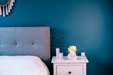 Photo for Cozy bedroom with navy blue walls, white bedside table with ceramic female body-shaped vase with flower and burning candles. Dark modern stylish female room. Self recovery place at home - Royalty Free Image