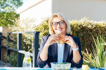 Téléchargez les photos : Portrait d'une femme d'affaires souriante d'âge moyen mangeant un hamburger sain pendant le déjeuner au café extérieur du parc pendant sa pause. Habitudes alimentaires saines et bien-être - en image libre de droit
