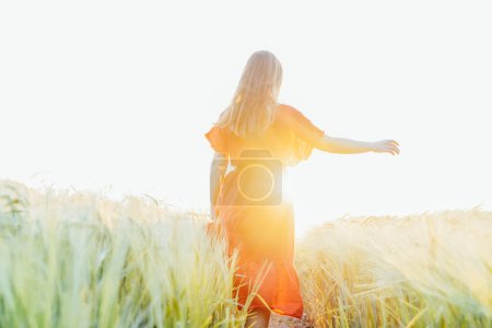 Rückenansicht Frau in rotem Kleid, die in einem Weizenfeld im Sonnenuntergang mit Sonnenstrahlen spazieren und entspannen geht. Ein einfaches Vergnügen für die psychische Gesundheit. Entspannung in der Natur. Sommerzeit. Weiche selektive Fokussierung. Kopierraum