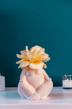 Close up white table with ceramic female body-shaped vase with rose flower, burning candles on turquoise blue wall background. Zen, cozy, meditation female bedroom decor. Self recovery place at home
