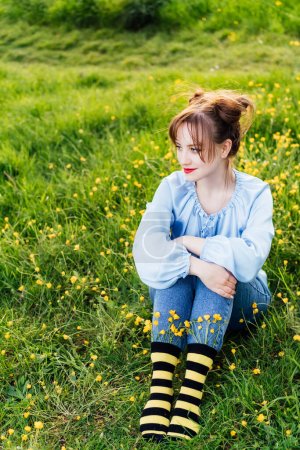 Junge Frau in blauer Bluse, Jeans und gestreiften schwarz-gelben Socken mit Blumen drinnen auf dem grünen Gras der blühenden Wiese sitzend. Anime-Stil. Konzept des Bienenschutzes, Blütezeit.