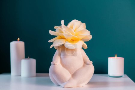 Close up white table with ceramic female body-shaped vase with rose flower, burning candles on turquoise blue wall background. Zen, cozy, meditation female bedroom decor. Self recovery place at home