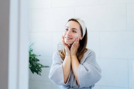 Young smiling woman applying cream mask and doing manual self massage of face in the bathroom. Natural procedures for skin care. Home Beauty treatment, face gymnastics for youth. Selective focus