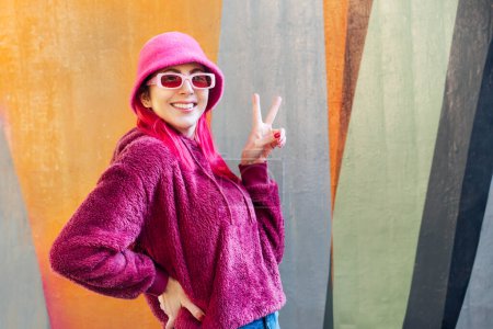 Urbane Herbst Winter Streetfashion. Kawaii-Stimmung. Lächelnde junge Frau mit rosa Haaren und Sonnenbrille im magentaflauschigen Sweatshirt und Eimerhut, die mit den Fingern V-Zeichen auf Graffiti-Hintergrund macht