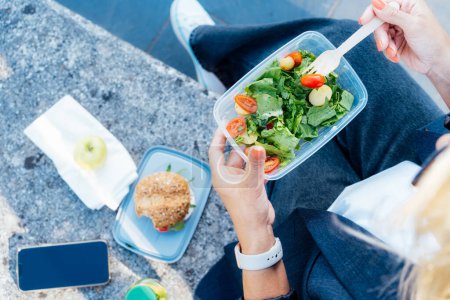 Téléchargez les photos : Top view close up business woman eating salad from her lunch box sitting on the bench at office park. Balanced diet lunchbox for weight loss. Healthy eating habits and well-being. Selective focus. - en image libre de droit