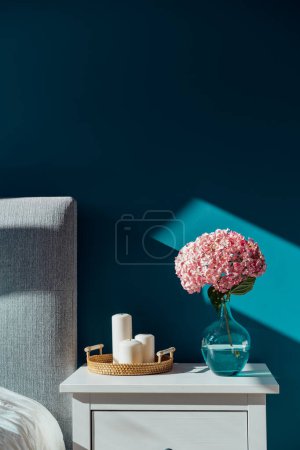Téléchargez les photos : Stylish modern cosy bedroom in dark colors. Cozy interior with turquoise walls, home decor. Bed with grey fabric headboard, white blanket, bedside table, vase with hydrangea flower, candles. Vertical - en image libre de droit