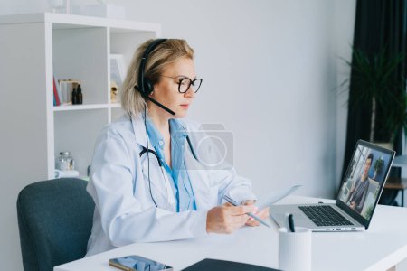Photo for Middle aged doctor in white coat and video conferencing headset make online video call consult patient on laptop. Telemedicine concept for domestic health treatment. Online remote medical appointment - Royalty Free Image