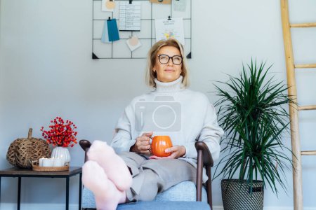 Middle aged woman relaxing with pumpkin shaped cup of hot drink in scandy style hygge interior home with fall mood decor. Lady dreaming, enjoy calm mood without stress, well being alone. Cozy autumn