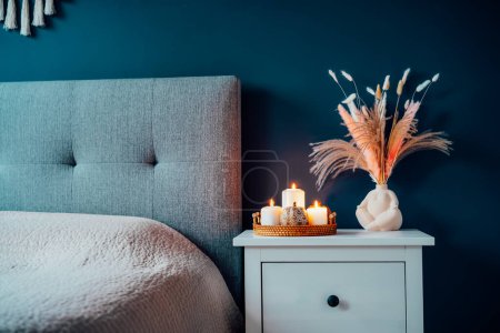 Autumn decor in stylish modern bedroom. Cozy interior with dark emerald wall, bed with grey fabric headboard, beige blanket, bedside table, vase with pampas grass, pumpkin and burning candles on tray